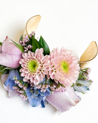 A wrist corsage arranged with fresh seasonal blooms and textural greenery, with decorative blue ribbon on a white background.