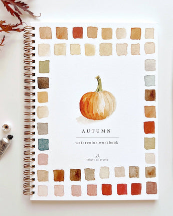 Watercolor workbook titled 'Autumn' with color swatches and a pumpkin illustration, surrounded by art supplies on a white background.
